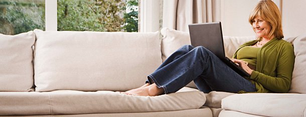Frau fühlt sich sicher bei der Partnersuche mit ElitePartner und sitzt zufrieden mit Laptop auf dem Schoß auf dem Sofa