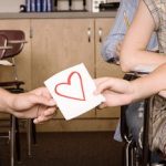 Symbolische Darstellung der ersten Liebe: männliche und weibliche Hand halten gemeinsam an Papier mit rotem Herz in Klassenzimmer