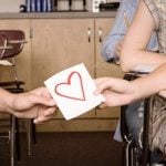 Symbolische Darstellung der ersten Liebe: männliche und weibliche Hand halten gemeinsam an Papier mit rotem Herz in Klassenzimmer